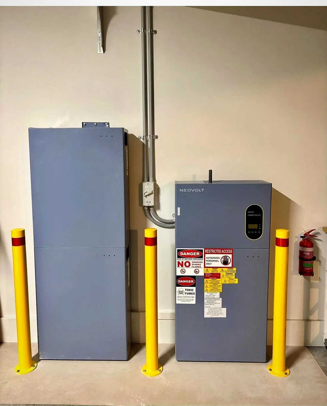 Two battery cabinets and an inverter on a garage wall with yellow bollards in front